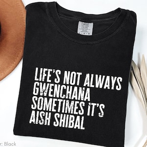 May include: Black t-shirt with the text "LIFE'S NOT ALWAYS GWENCHANA SOMETIMES IT'S AISH SHIBAL" in white block letters. The shirt is folded and has a Comfort Colors tag. A brown hat and dried plant are in the background.