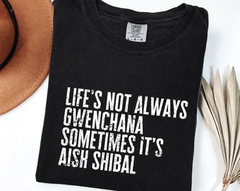 Puede incluir: Camiseta negra con el texto "LIFE'S NOT ALWAYS GWENCHANA SOMETIMES IT'S AISH SHIBAL" en letras blancas. La camiseta está doblada y tiene una etiqueta Comfort Colors. Un sombrero marrón y una planta seca están en el fondo.