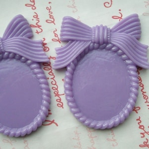 May include: Two lavender resin oval embellishments, each with a bow at the top. The oval frames have a rope-like border. The bows have a ribbed texture. The background is a light-coloured paper with handwritten text.