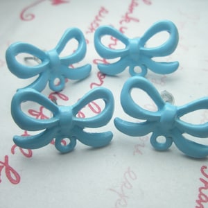 May include: Four light blue bow-shaped stud earrings. The earrings are made of a solid material and have a glossy finish. The bows are arranged in a cluster on a patterned white surface. The earrings are a cute accessory.