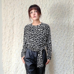 Op de afbeelding: Een blouse met lange mouwen met een zwart-wit luipaardprint en een strikdetail in de taille, gecombineerd met een zwarte broek met hoge taille en zwarte schoenen met een puntige neus. De blouse heeft een ronde halslijn.
