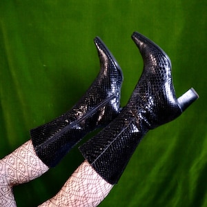 Puede incluir: Un par de botas negras hasta la rodilla con un estampado de piel de serpiente. Las botas tienen puntas puntiagudas y tacones pequeños. El fondo es de color verde oscuro.