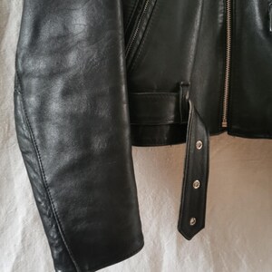 K&ouml;nnte beinhalten: Schwarze Lederjacke mit Rei&szlig;verschluss und G&uuml;rtel in der Taille. Die Jacke hat einen langen &Auml;rmel und einen spitzen Kragen.