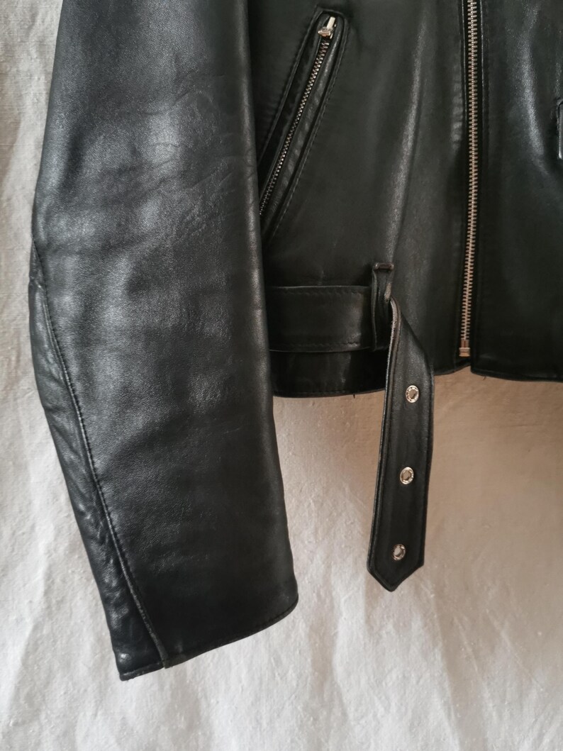K&ouml;nnte beinhalten: Schwarze Lederjacke mit Rei&szlig;verschluss und G&uuml;rtel in der Taille. Die Jacke hat einen langen &Auml;rmel und einen spitzen Kragen.