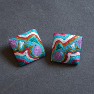 May include: Two square, embroidered earrings with a colorful, wavy pattern. The design includes teal, white, red, gold, and purple threads. The earrings are set against a gray background.