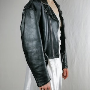 K&ouml;nnte beinhalten: Eine schwarze Lederjacke im Motorrad-Stil mit Rei&szlig;verschluss und silbernem Rei&szlig;verschluss-Zipper. Die Jacke hat eine verk&uuml;rzte L&auml;nge und eine leicht &uuml;berdimensionierte Passform.
