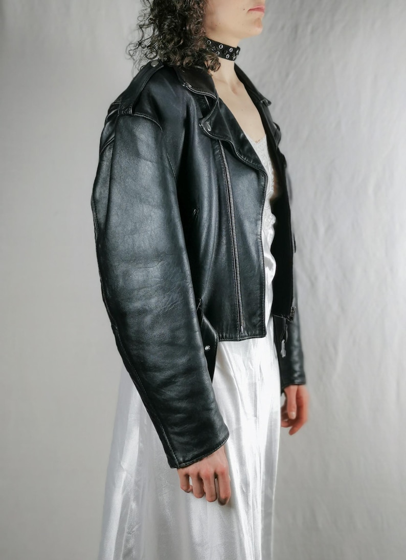 K&ouml;nnte beinhalten: Eine schwarze Lederjacke im Motorrad-Stil mit Rei&szlig;verschluss und silbernem Rei&szlig;verschluss-Zipper. Die Jacke hat eine verk&uuml;rzte L&auml;nge und eine leicht &uuml;berdimensionierte Passform.