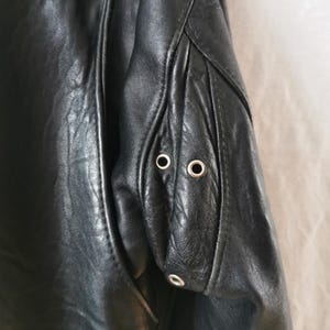 K&ouml;nnte beinhalten: Schwarze Lederjacke mit Rei&szlig;verschluss und zwei silbernen &Ouml;sen am &Auml;rmel.