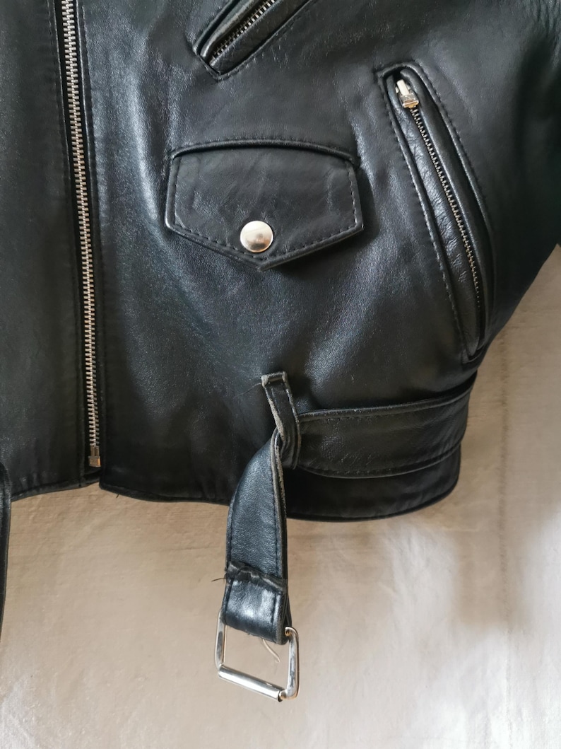 K&ouml;nnte beinhalten: Schwarze Lederjacke mit Rei&szlig;verschluss, einer Druckknopftasche und einem G&uuml;rtel mit einer silbernen Schnalle.