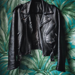 K&ouml;nnte beinhalten: Eine schwarze Lederjacke im Motorrad-Stil mit Rei&szlig;verschluss und mehreren Taschen. Die Jacke h&auml;ngt an einem Kleiderb&uuml;gel vor einem Hintergrund aus gr&uuml;nen und blauen tropischen Bl&auml;ttern.