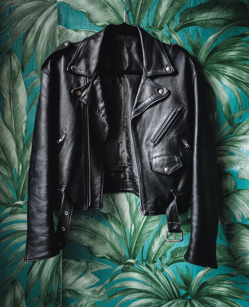 K&ouml;nnte beinhalten: Eine schwarze Lederjacke im Motorrad-Stil mit Rei&szlig;verschluss und mehreren Taschen. Die Jacke h&auml;ngt an einem Kleiderb&uuml;gel vor einem Hintergrund aus gr&uuml;nen und blauen tropischen Bl&auml;ttern.
