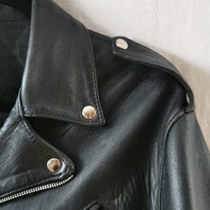 K&ouml;nnte beinhalten: Nahaufnahme einer schwarzen Lederjacke mit silbernen Druckkn&ouml;pfen und einem Rei&szlig;verschluss. Die Jacke hat einen klassischen Biker-Stil.