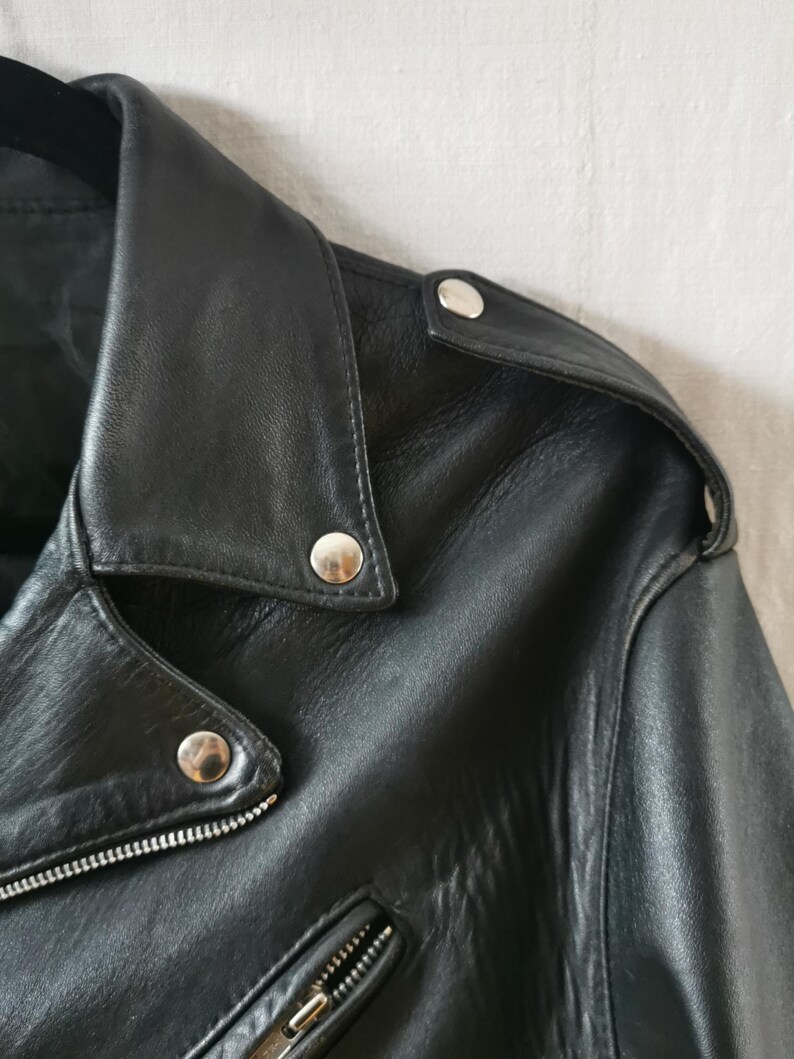K&ouml;nnte beinhalten: Nahaufnahme einer schwarzen Lederjacke mit silbernen Druckkn&ouml;pfen und einem Rei&szlig;verschluss. Die Jacke hat einen klassischen Biker-Stil.