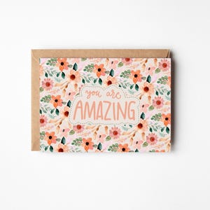May include: A floral greeting card with a kraft paper envelope. The card features a watercolor design of peach and pink flowers with green leaves. The words "you are AMAZING" are in a cloud-shaped text box.
