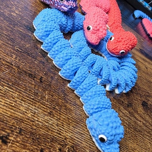 Puede incluir: Tres juguetes de serpiente articulados con cuerpos texturizados y segmentados. Una serpiente es azul, otra roja y una presenta un degradado de morado, azul y verde. Cada serpiente tiene una cabeza con ojos negros y blancos, dispuestas en espiral.