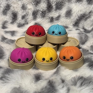 May include: Five colorful, round, squishy dumpling toys, each featuring a smiling face with black eyes and pink cheeks. Each dumpling sits inside a small, tan, circular bamboo-style steamer basket. The colors include red, blue, purple, yellow, and orange.