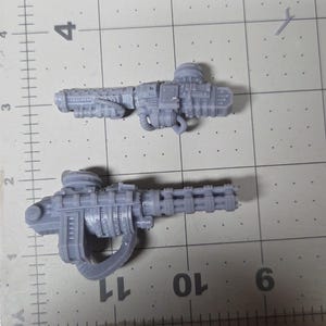May include: Two unpainted grey resin miniature weapon components featuring intricate mechanical details, including barrels, vents, and cables. The items are positioned on a cutting mat with visible grid lines, showing the top piece is 2.5 inches long.