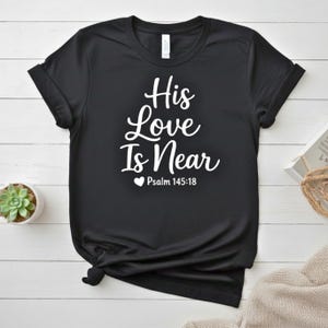 K&ouml;nnte beinhalten: Schwarzes T-Shirt mit dem wei&szlig;en Text "His Love Is Near" und "Psalm 145:18" unter einem Herzsymbol. Das Shirt ist gefaltet, mit einer kleinen Sukkulentenpflanze in einem wei&szlig;en Topf im Hintergrund.