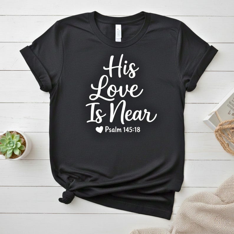 K&ouml;nnte beinhalten: Schwarzes T-Shirt mit dem wei&szlig;en Text "His Love Is Near" und "Psalm 145:18" unter einem Herzsymbol. Das Shirt ist gefaltet, mit einer kleinen Sukkulentenpflanze in einem wei&szlig;en Topf im Hintergrund.