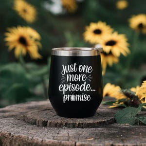 May include: A black stainless steel tumbler with a silver rim. The tumbler has white text that reads "just one more episode... promise" with a television graphic. The tumbler is sitting on a wooden surface with yellow flowers in the background.