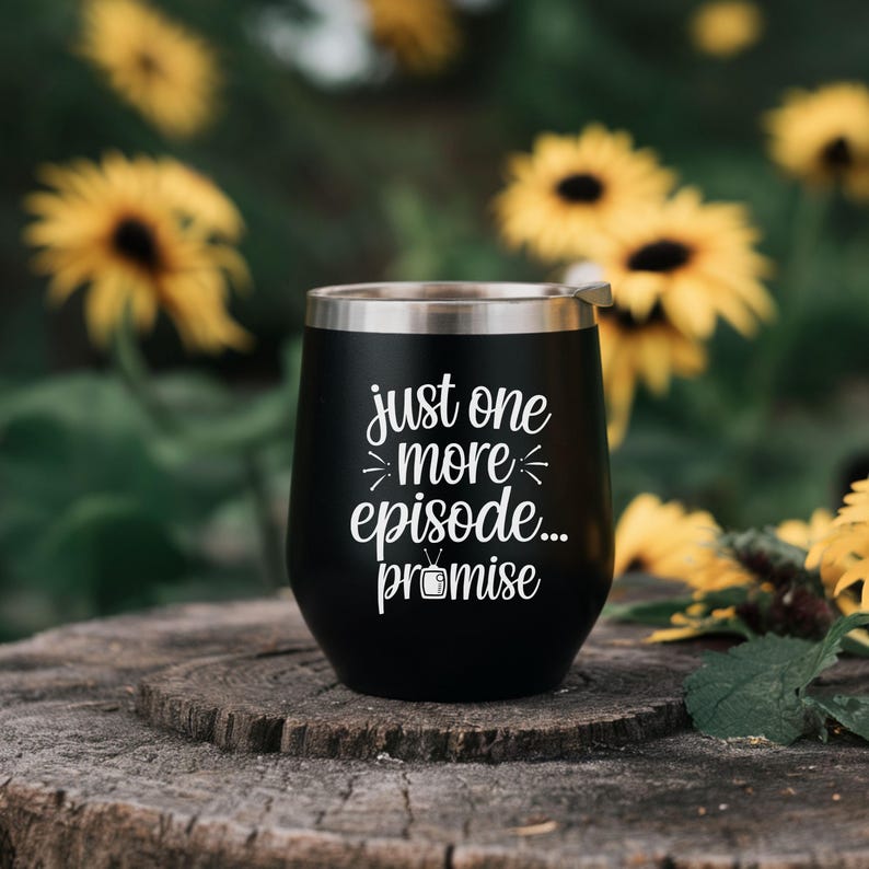 May include: A black stainless steel tumbler with a silver rim. The tumbler has white text that reads "just one more episode... promise" with a television graphic. The tumbler is sitting on a wooden surface with yellow flowers in the background.