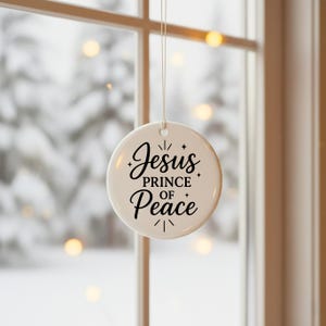 May include: A round, white Christmas ornament with the words "Jesus Prince of Peace" in black script. The ornament is hanging in front of a window with a blurred winter scene visible in the background.