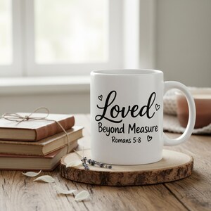 K&ouml;nnte beinhalten: Wei&szlig;e Keramiktasse mit den Worten "Loved Beyond Measure" und "Romans 5:8" in schwarzer Schrift. Die Tasse steht auf einem Holzuntersetzer, mit B&uuml;chern und dekorativen Elementen im Hintergrund.