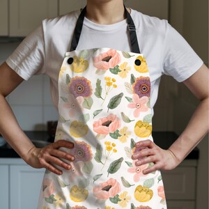 K&ouml;nnte beinhalten: Eine wei&szlig;e Sch&uuml;rze mit einem Blumenmuster mit rosa, gelben und lila Blumen und gr&uuml;nen Bl&auml;ttern. Die Sch&uuml;rze hat einen schwarzen Nackenriemen und schwarze Kn&ouml;pfe. Die Sch&uuml;rze wird von einer Person getragen.
