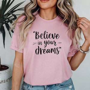 May include: A pale pink t-shirt featuring the black text "Believe in your dreams". The short-sleeved top has a round neck. The slogan is printed centrally on the front of the garment.