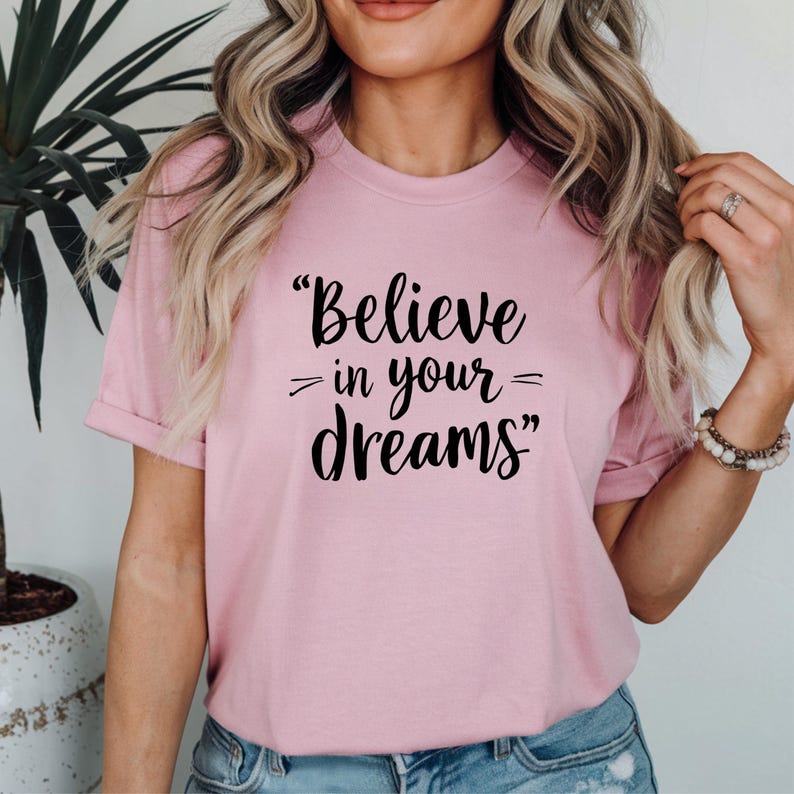 May include: A pale pink t-shirt featuring the black text "Believe in your dreams". The short-sleeved top has a round neck. The slogan is printed centrally on the front of the garment.