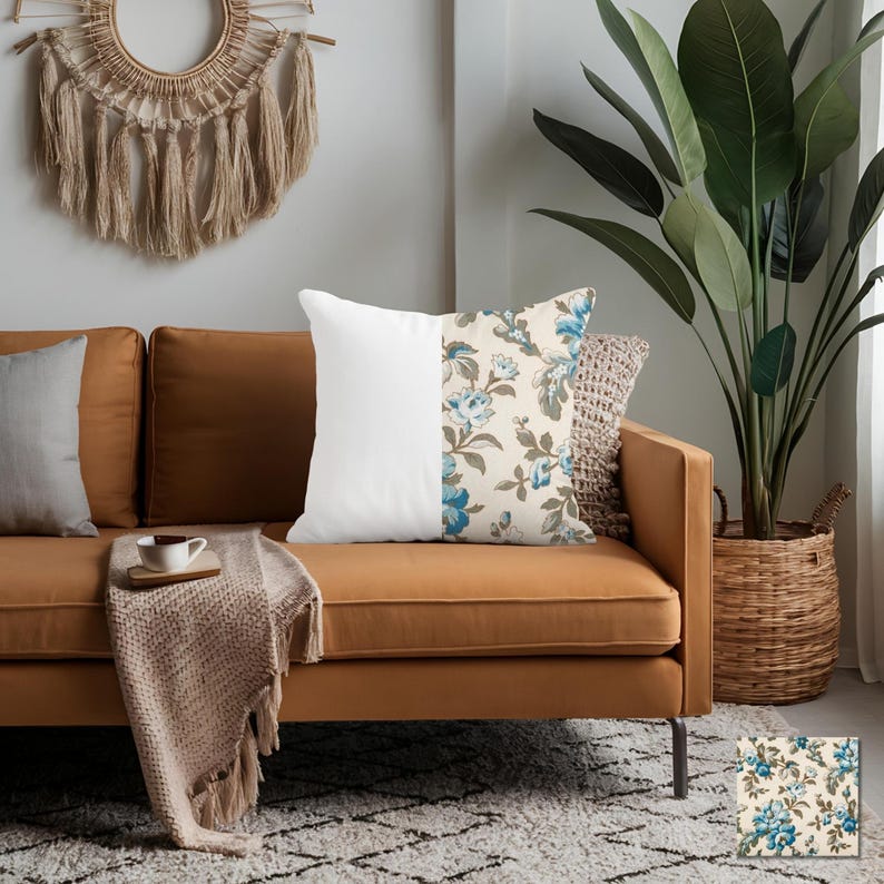 May include: A decorative throw pillow with a split design. One half is solid white, the other features a floral pattern with blue flowers and green leaves on a cream background. The pillow is placed on a brown sofa.