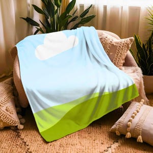 Puede incluir: Una manta azul suave con un diseño de nube blanca y paisaje verde. La manta está sobre una silla, creando un ambiente acogedor y acogedor. El diseño evoca una sensación de naturaleza y tranquilidad.