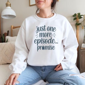 May include: White sweatshirt with the text "just one more episode... promise" in dark teal. The sweatshirt is a crew neck style and is paired with blue jeans. The text includes a small television graphic.
