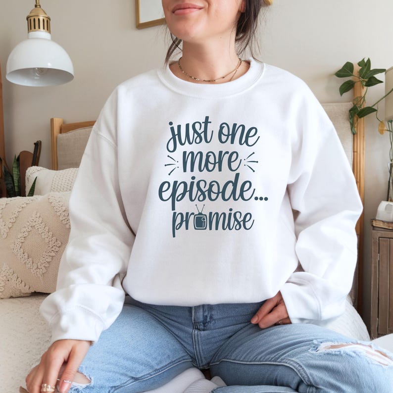 May include: White sweatshirt with the text "just one more episode... promise" in dark teal. The sweatshirt is a crew neck style and is paired with blue jeans. The text includes a small television graphic.