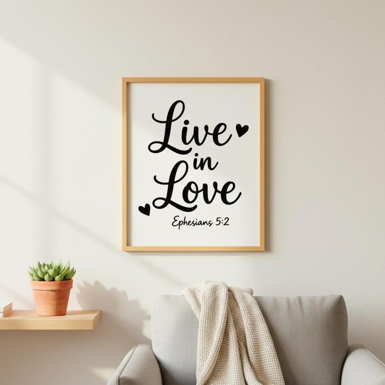 K&ouml;nnte beinhalten: Ein gerahmter Druck mit dem Schriftzug "Live in Love" in schwarzer Schrift, verziert mit Herzen. Der Text "Ephesians 5:2" steht darunter. Der Rahmen ist hellbraun. Das Bild h&auml;ngt an einer wei&szlig;en Wand.