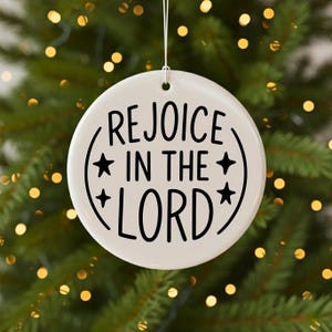 May include: A white, round Christmas ornament with the words "REJOICE IN THE LORD" in black script. The text is surrounded by a curved line and stars. The ornament hangs from a white string.