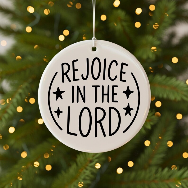 May include: A white, round Christmas ornament with the words "REJOICE IN THE LORD" in black script. The text is surrounded by a curved line and stars. The ornament hangs from a white string.