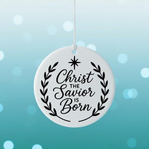May include: A white, round Christmas ornament with the text "Christ the Savior is Born" in elegant script, surrounded by a black laurel wreath. A black star is above the text. The ornament hangs from a white string.