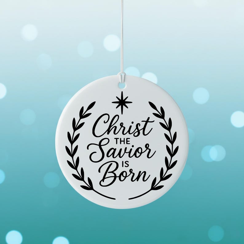 May include: A white, round Christmas ornament with the text "Christ the Savior is Born" in elegant script, surrounded by a black laurel wreath. A black star is above the text. The ornament hangs from a white string.