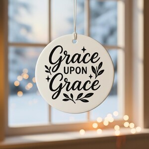 May include: A round, off-white ornament with the words "Grace upon Grace" in elegant black script. The design includes decorative flourishes and is suspended by a silver string. The background shows a window with a blurred winter scene.