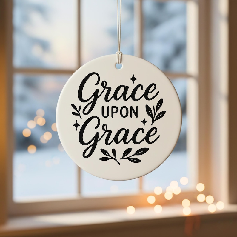 May include: A round, off-white ornament with the words "Grace upon Grace" in elegant black script. The design includes decorative flourishes and is suspended by a silver string. The background shows a window with a blurred winter scene.