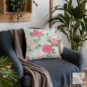 May include: Square throw pillow with a floral pattern of pink and white roses on a light blue background. The pillow is placed on a dark blue armchair with a brown blanket.