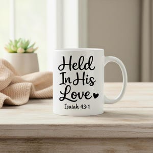 K&ouml;nnte beinhalten: Wei&szlig;e Keramik-Tasse mit dem Text "Held In His Love" in schwarzer Schrift, zusammen mit "Isaiah 43:1". Die Tasse hat einen gebogenen Henkel und steht auf einer Holzoberfl&auml;che.