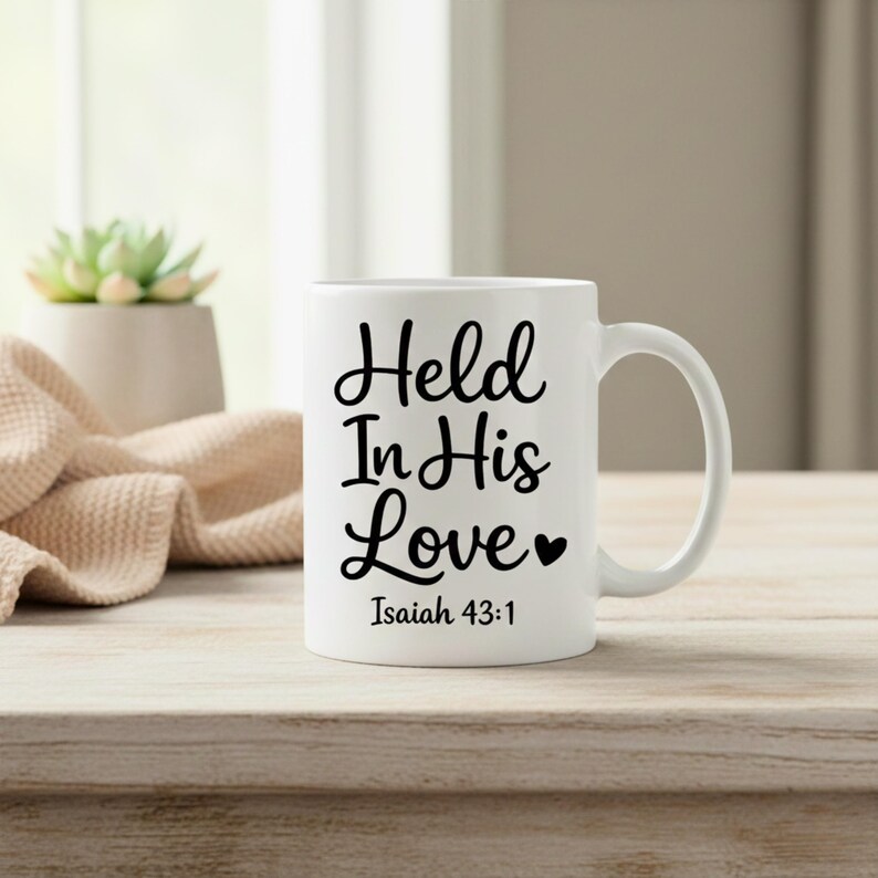 K&ouml;nnte beinhalten: Wei&szlig;e Keramik-Tasse mit dem Text "Held In His Love" in schwarzer Schrift, zusammen mit "Isaiah 43:1". Die Tasse hat einen gebogenen Henkel und steht auf einer Holzoberfl&auml;che.
