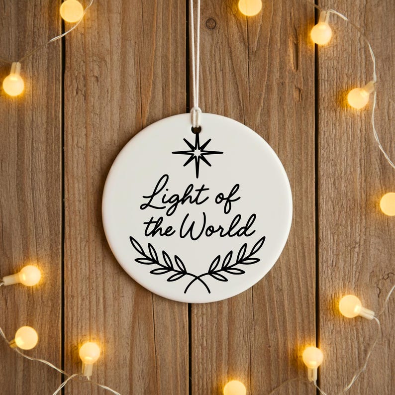 May include: A white, round ceramic ornament with the text "Light of the World" in black script, adorned with a star and leaf design. The ornament hangs from a white string against a wooden background, surrounded by glowing string lights.