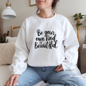 May include: A white crew neck sweatshirt featuring the black script text "Be your own kind of Beautiful". The sweatshirt is worn with blue jeans. The overall style is casual and comfortable.