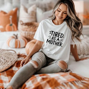 May include: White t-shirt with the text "TIRED. AS A MOTHER." in black. The shirt is paired with distressed gray jeans. The background includes pumpkins and fall-themed decor.