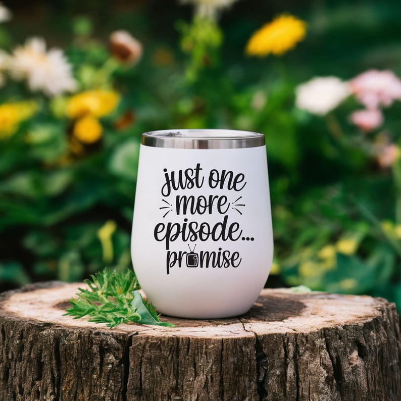 May include: A white insulated wine tumbler with a silver rim. The tumbler has the phrase "just one more episode... promise" in black script. The tumbler is sitting on a tree stump with a blurred green and yellow background.