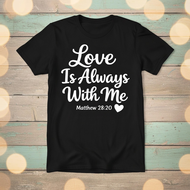 K&ouml;nnte beinhalten: Schwarzes T-Shirt mit dem wei&szlig;en Text "Love Is Always With Me" und dem Verweis "Matth&auml;us 28,20" mit einem Herzsymbol. Das Shirt wird vor einem hellen Holzmaserungshintergrund gezeigt.