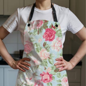 K&ouml;nnte beinhalten: Sch&uuml;rze mit floralem Muster mit rosa und pfirsichfarbenen Rosen auf hellblauem Hintergrund, erg&auml;nzt durch gr&uuml;nes Laub. Sie hat schwarze Tr&auml;ger und Kn&ouml;pfe. Die Person tr&auml;gt ein wei&szlig;es T-Shirt.